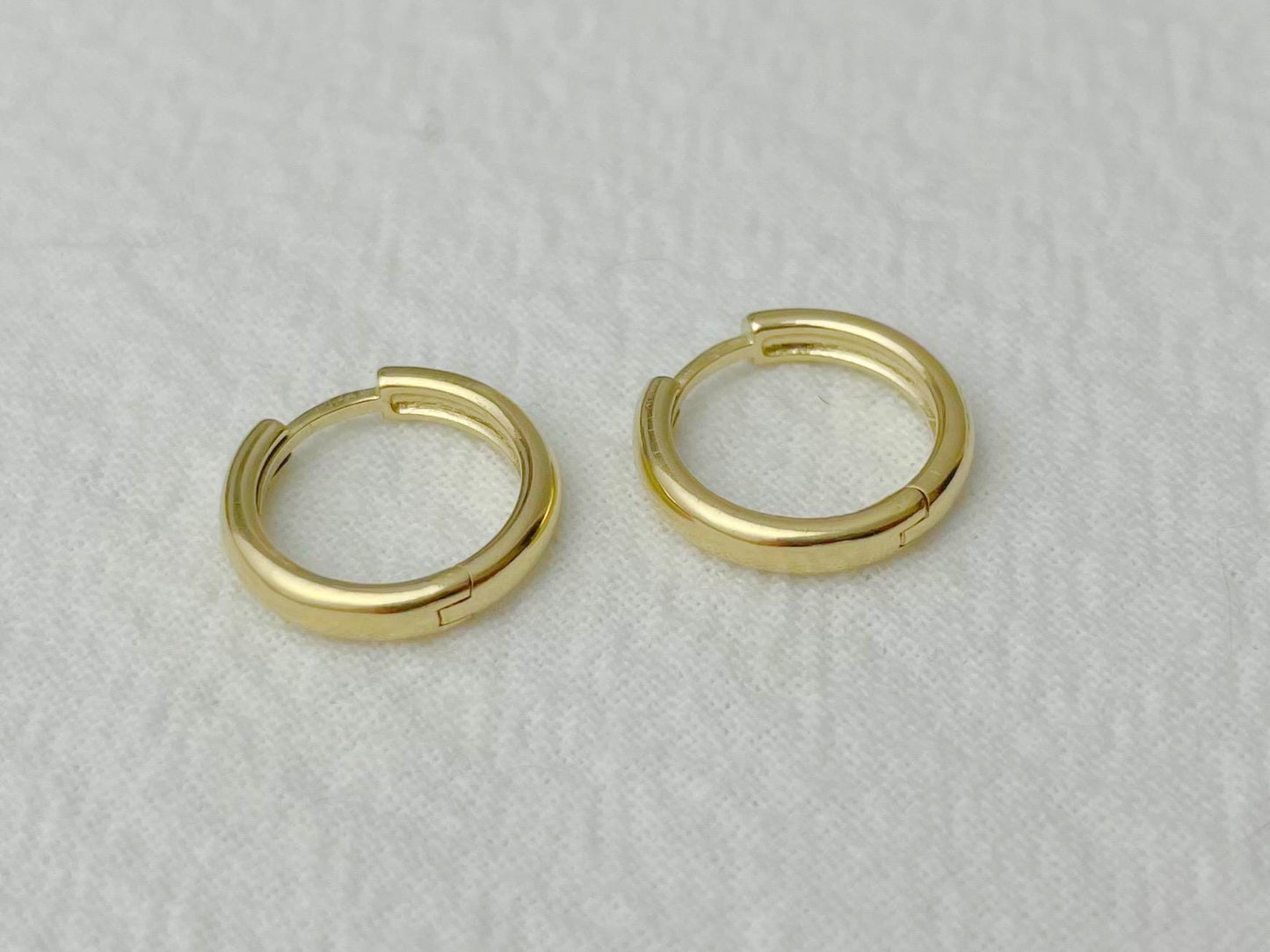 Petite yellow gold hoops reflecting soft light, photographed flat on a white cloth
